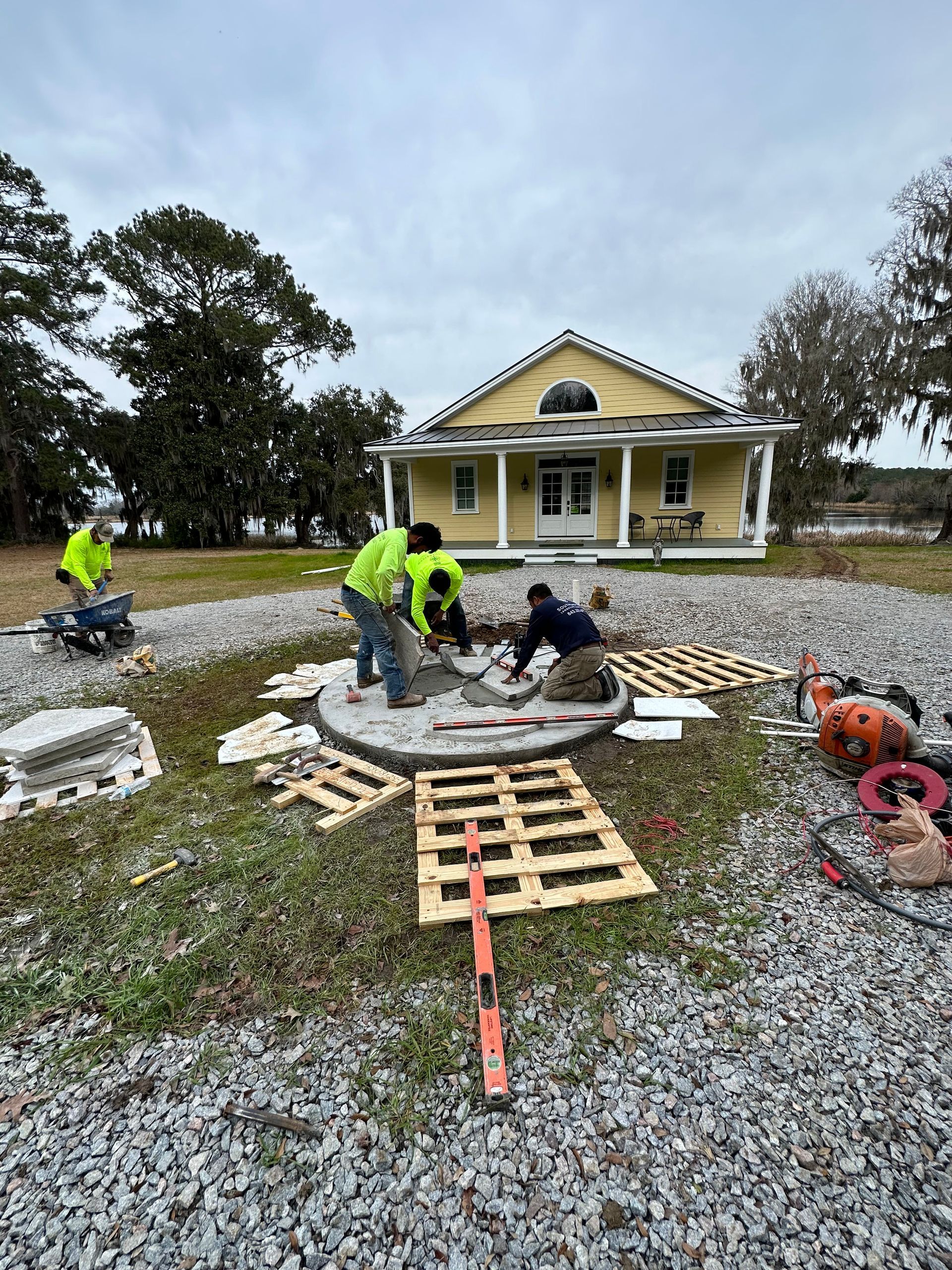 Construction workers installing a brick patio in front of a yellow house. Cloudy sky, gravel ground.