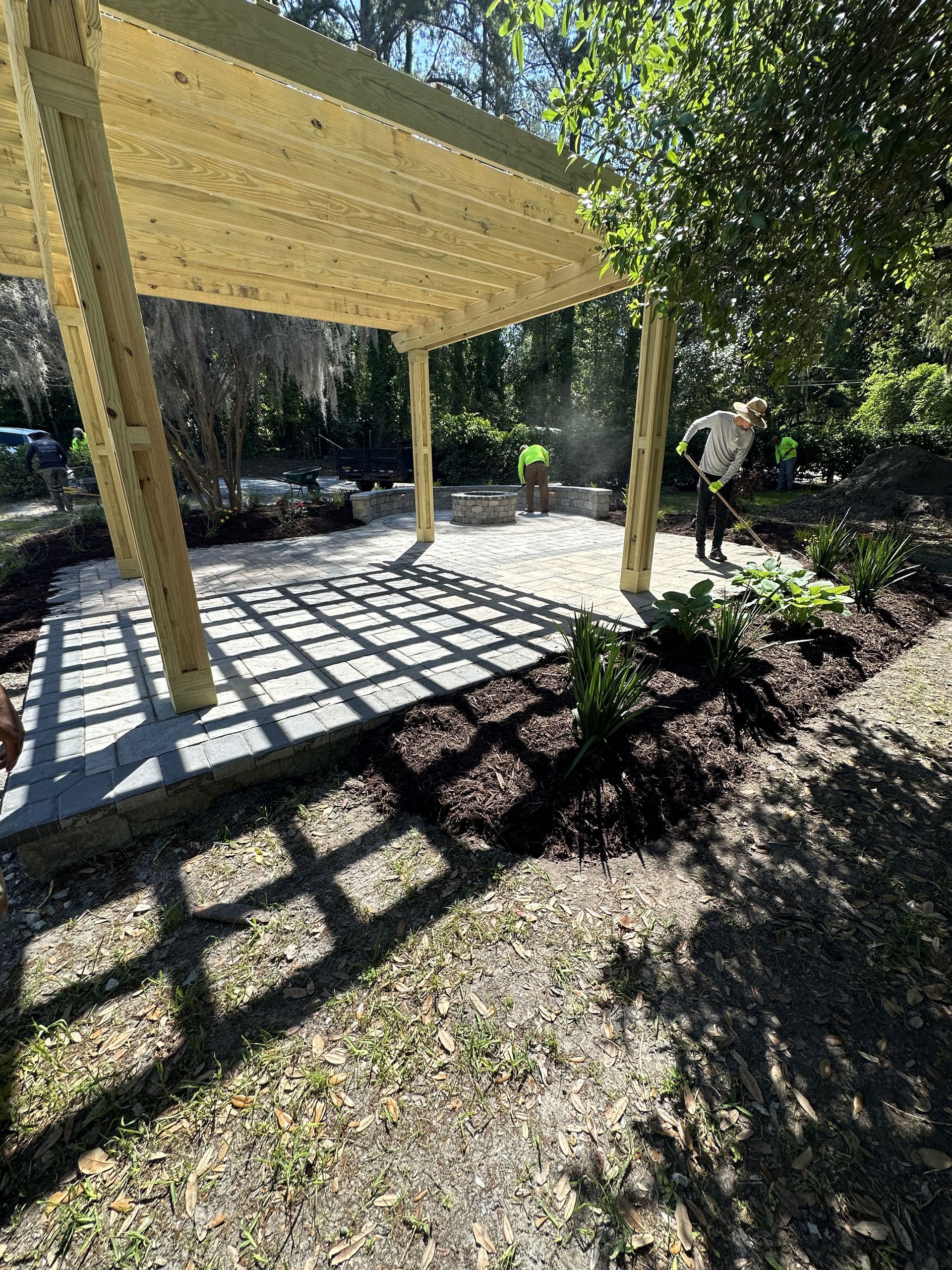 Pergola with gridded roof casts a shadow on a stone patio with person gardening.