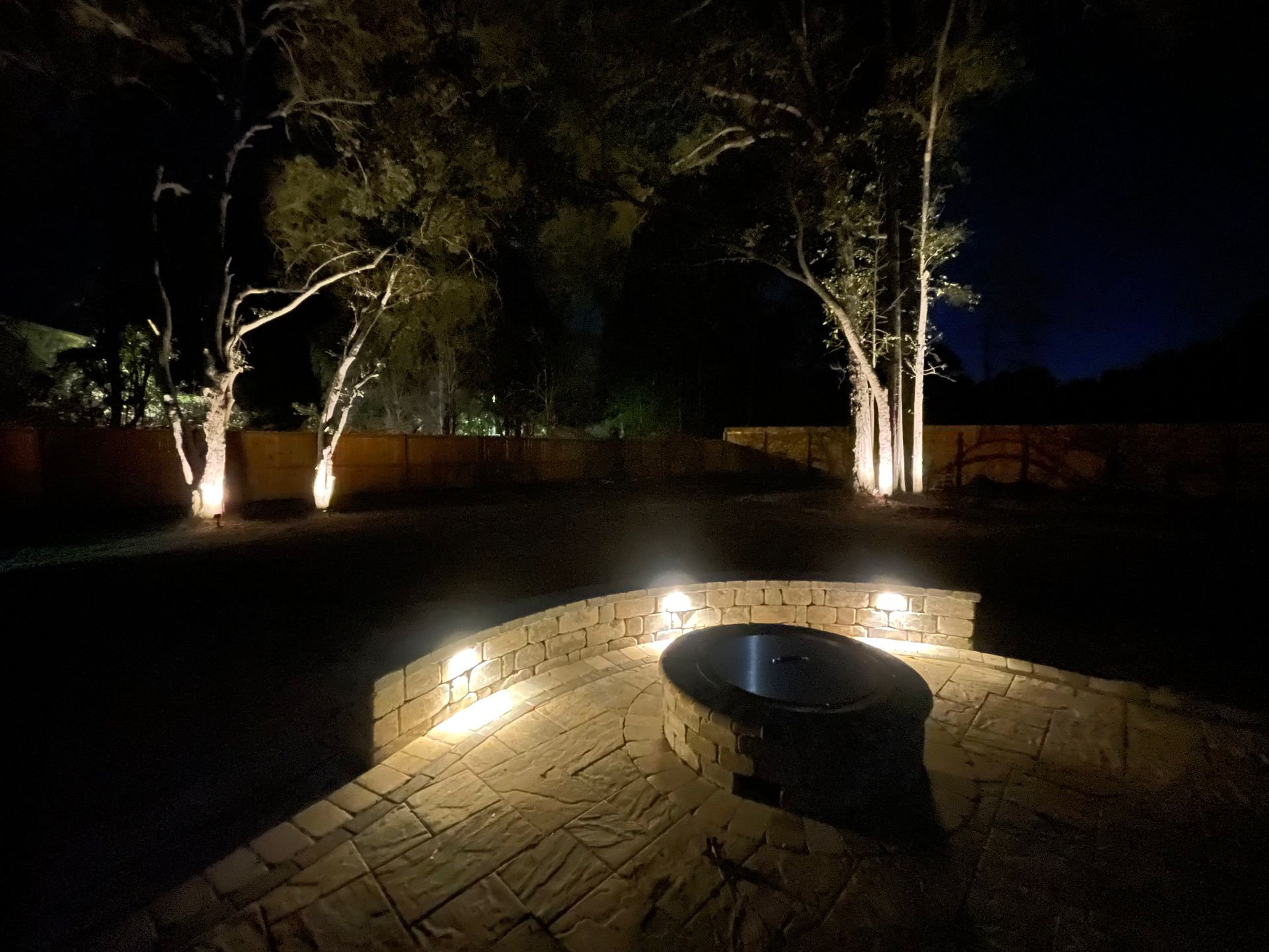 Night scene: backyard with trees and a fire pit illuminated by landscape lighting.