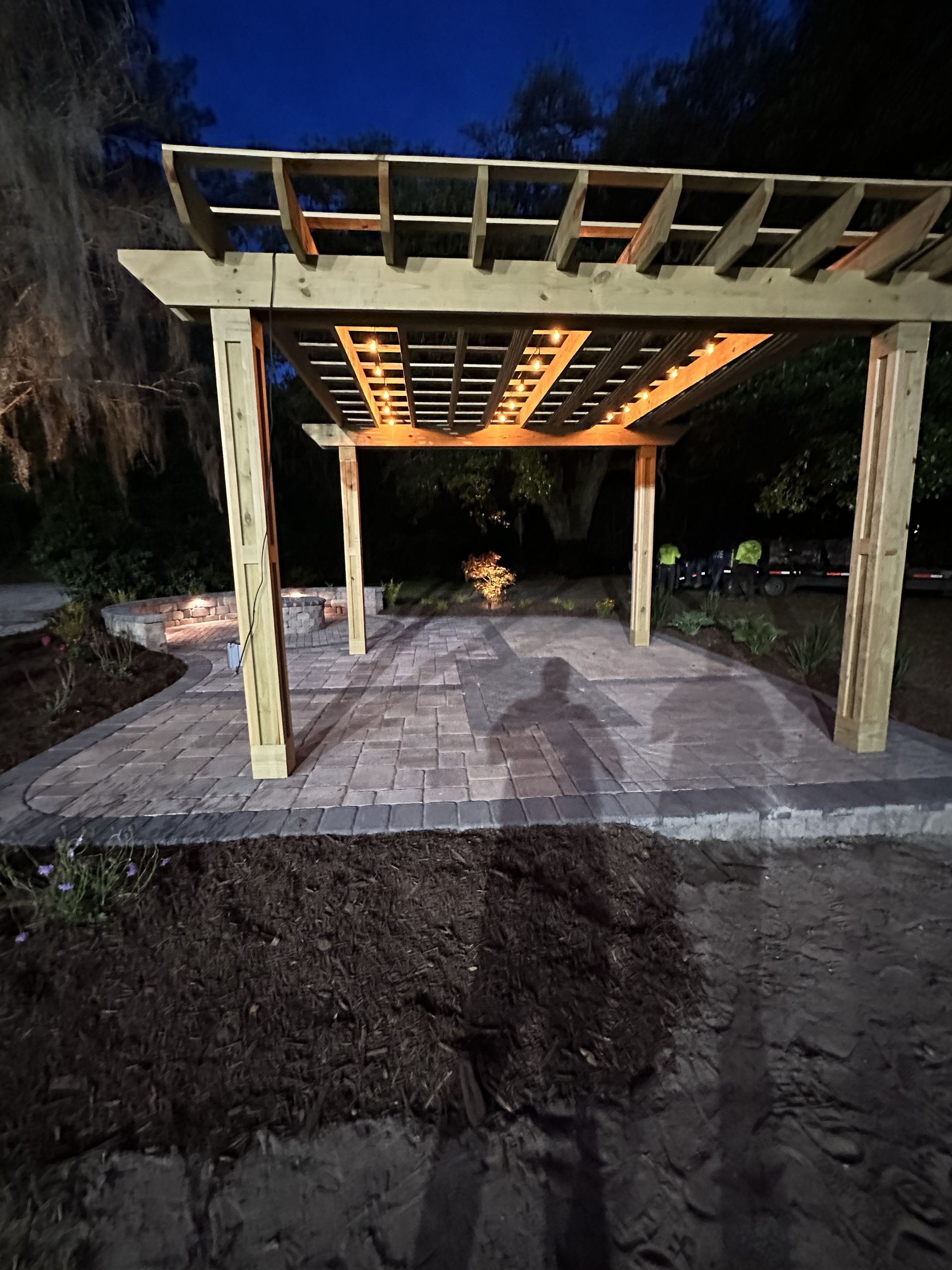 Wooden pergola over a brick patio, at dusk. Shadow of person on patio floor.