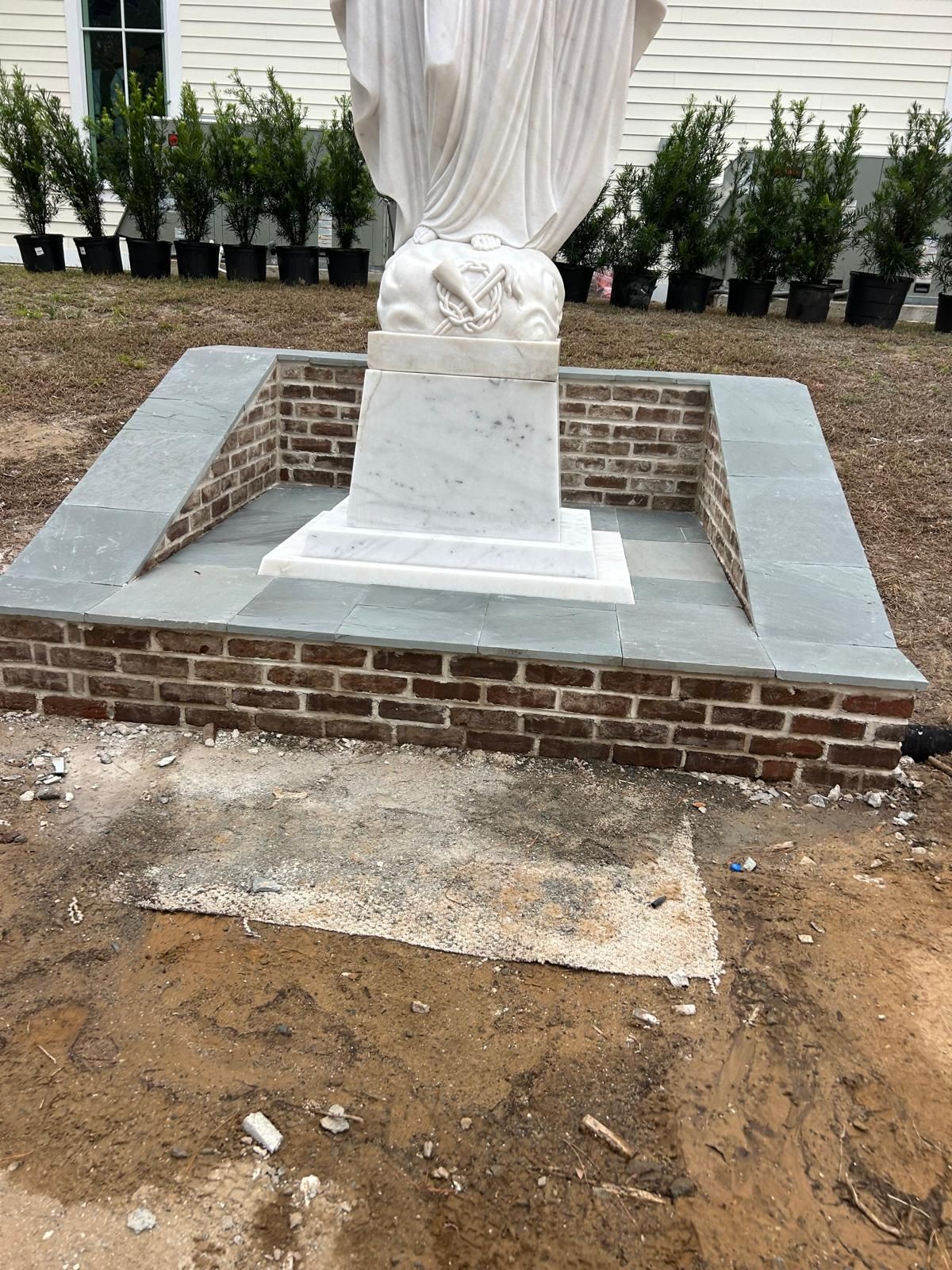 Statue on brick and stone pedestal; plants in pots behind it; dirt and construction site.