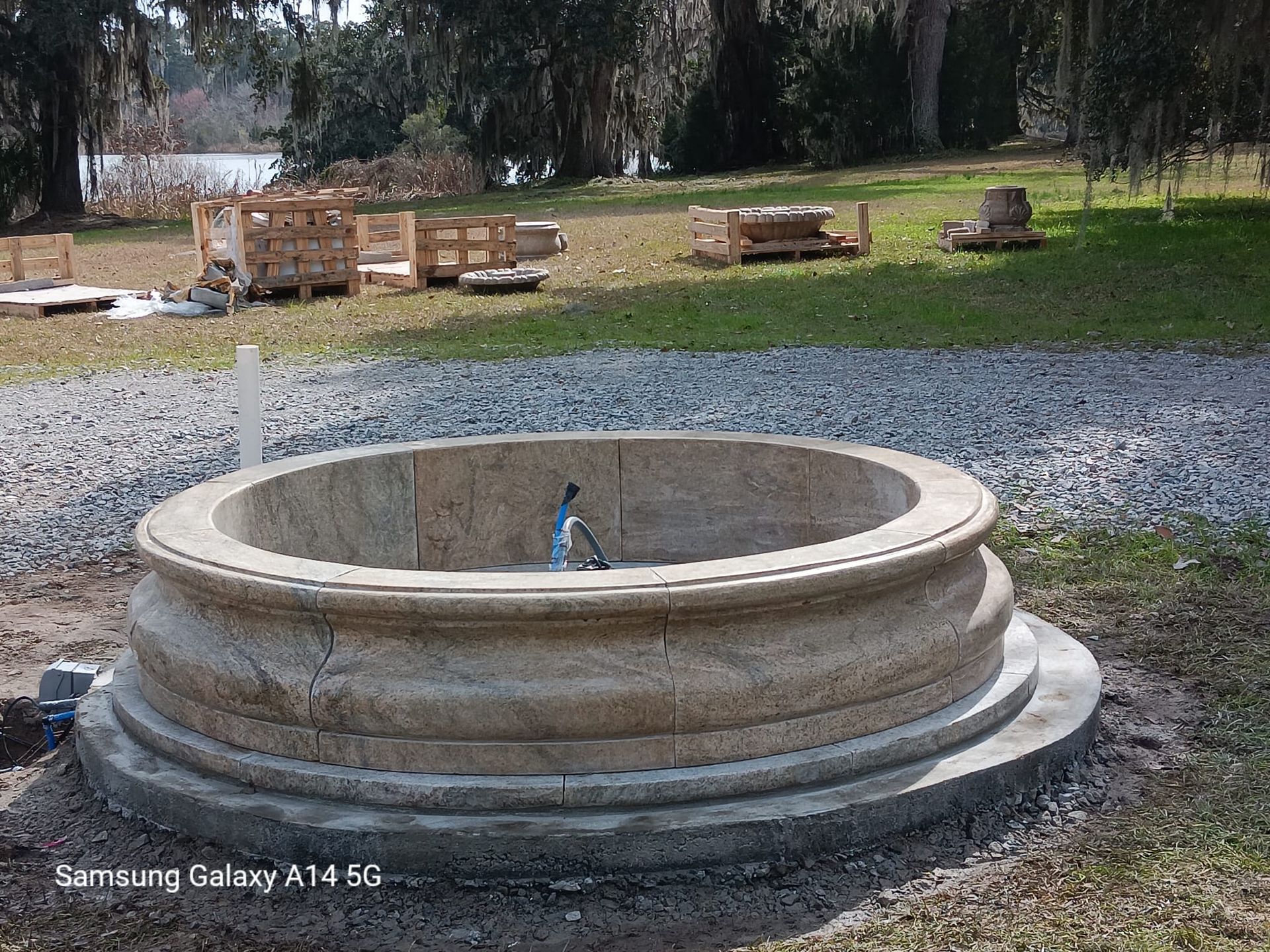 Stone well with ornate rim, outdoors on a grassy area near a lake. Construction materials in the background.