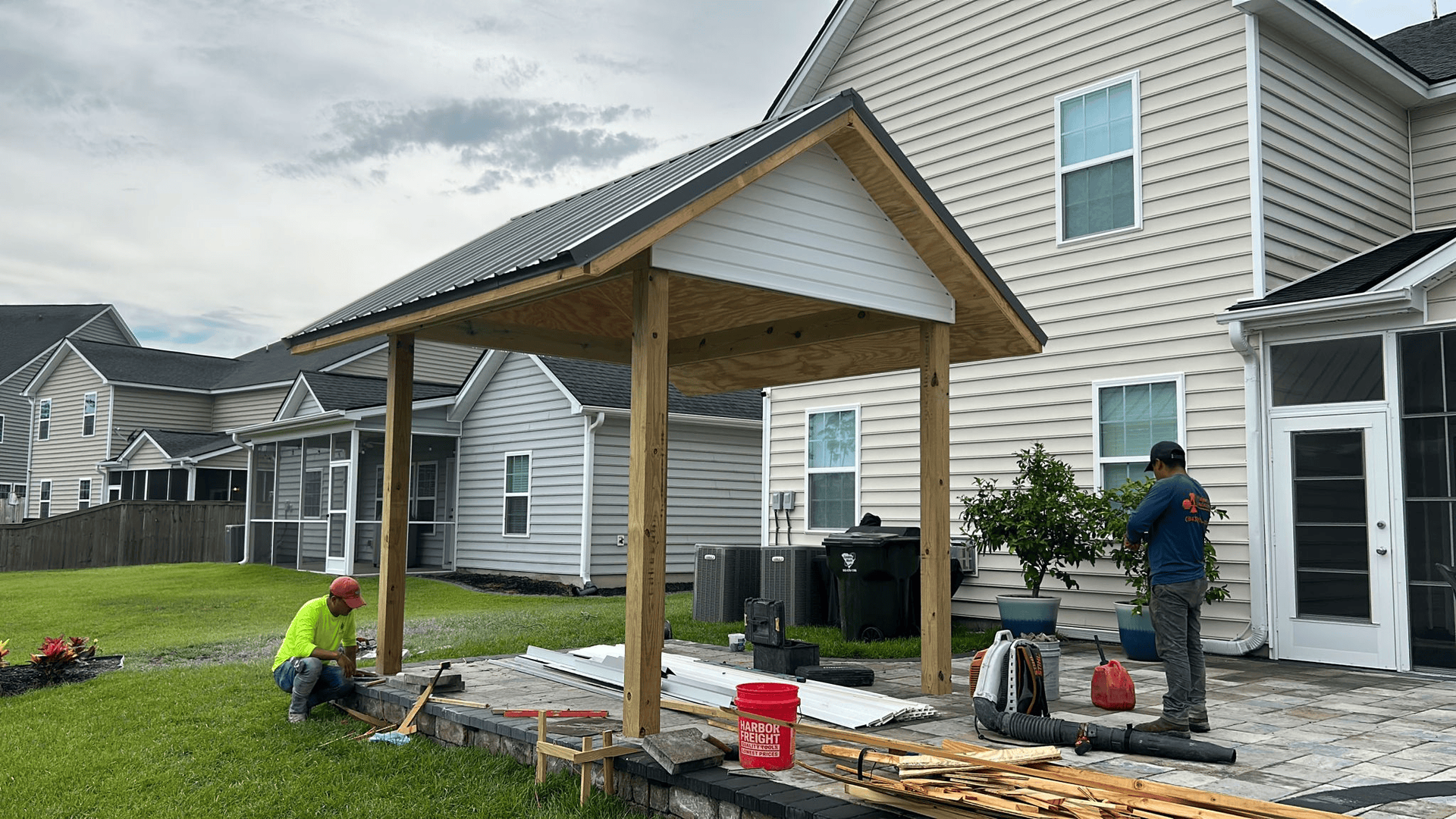 Construction of a backyard canopy; workers on patio and grass, building structure with wooden beams and a corrugated metal roof.