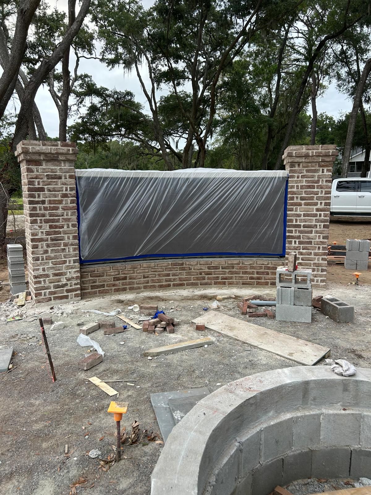 Brick wall with pillars under construction, gray tarp covering a central area, surrounded by tools and materials outdoors.