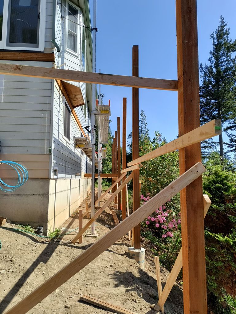 Wooden construction framing beside a house, with diagonal braces and a dirt path along the foundation.