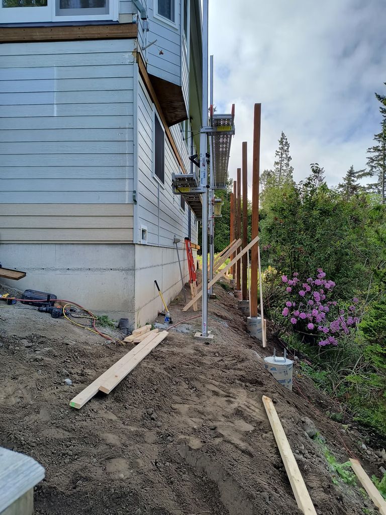 Construction work beside house with posts, muddy ground, and a pink flowering bush