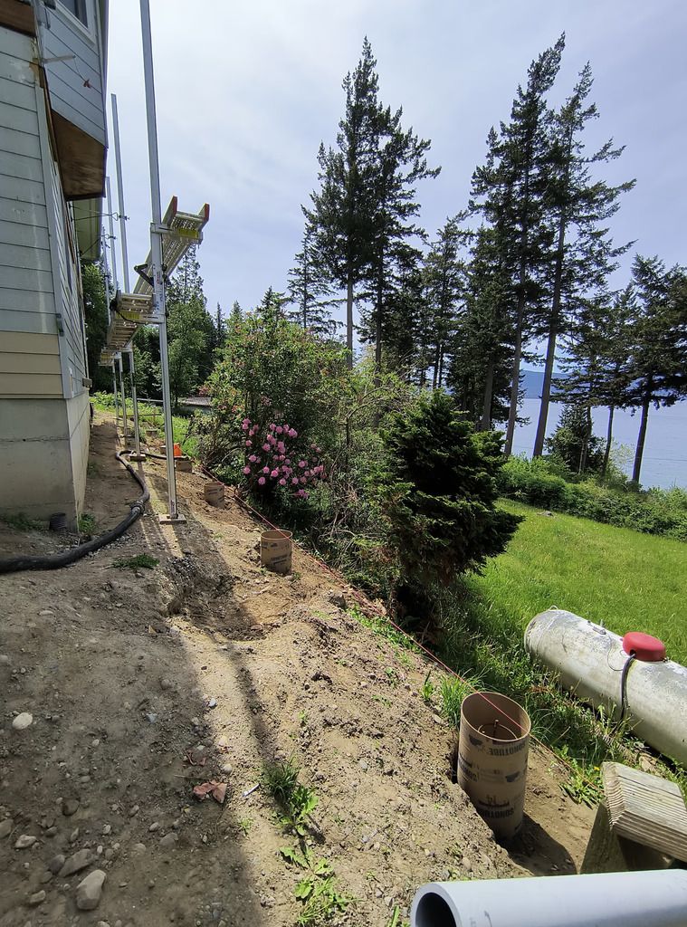 Narrow garden path beside a house with shrubs, grass, and tall trees overlooking water.
