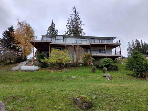 A house with a covered deck sits on a grassy hillside under a cloudy sky.