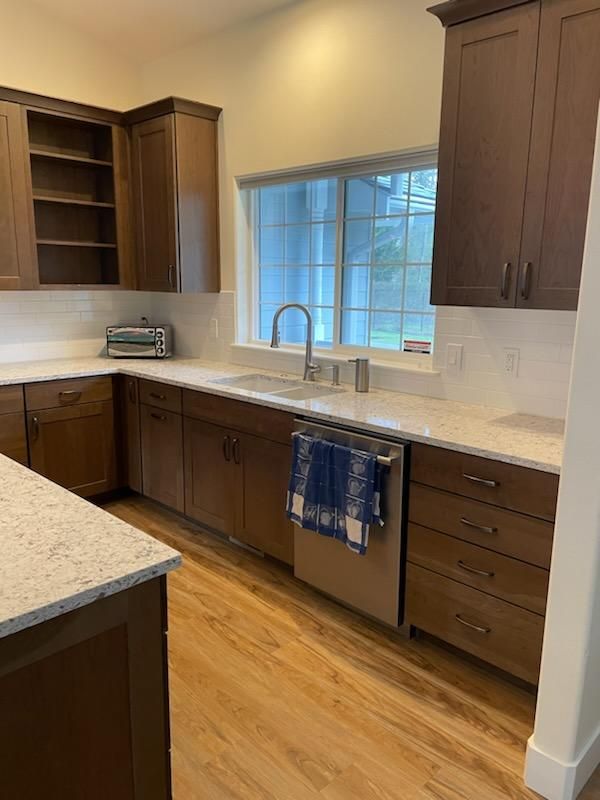 Empty modern kitchen with wood cabinets, light counters, stainless dishwasher, and a large window over the sink