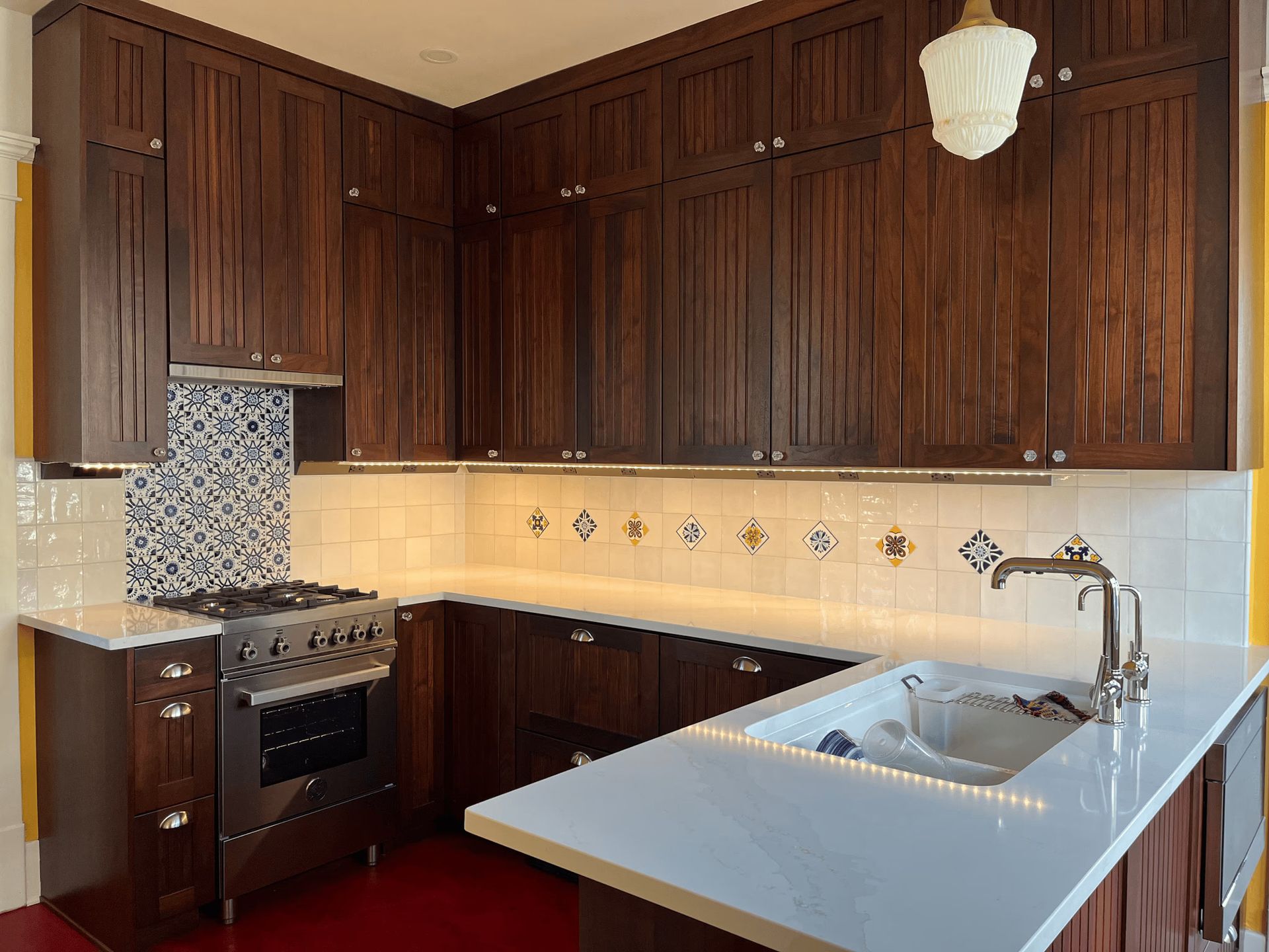 Compact kitchen with dark wood cabinets, white countertops, tiled backsplash, stove, sink, and hanging light fixture