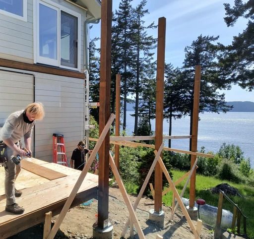 Person working on a lakeside deck frame beside a house, with wooden posts and tools in a sunny yard.