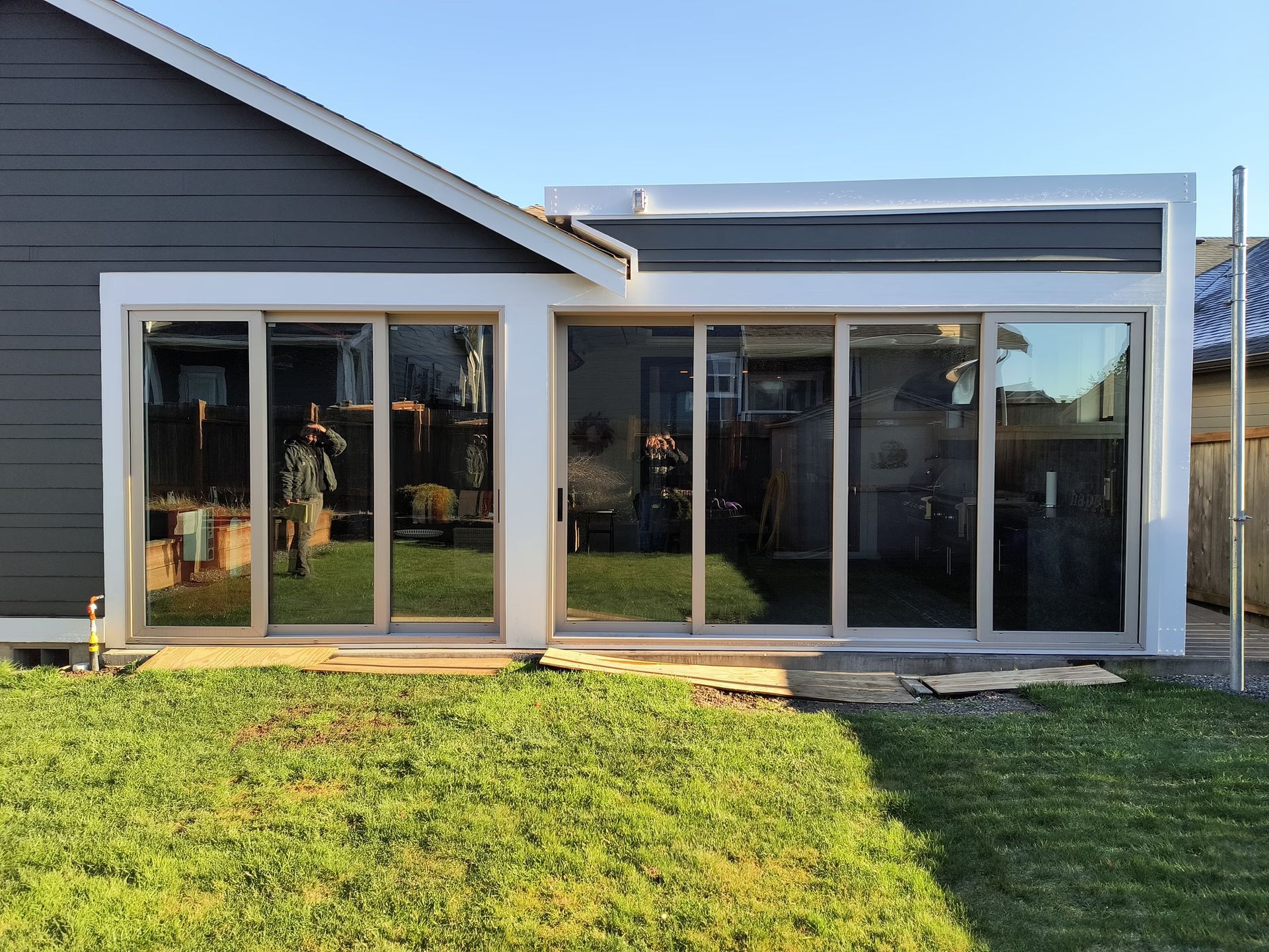 Glass sunroom attached to a gray house, with sliding doors opening to a grassy backyard.