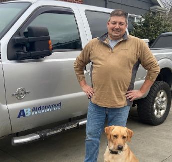 Man standing beside a silver service truck with a yellow dog in front, outdoors on a driveway