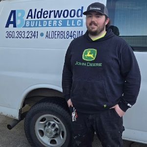 Man standing beside an Alderwood Builders LLC van, wearing a John Deere hoodie and cap.