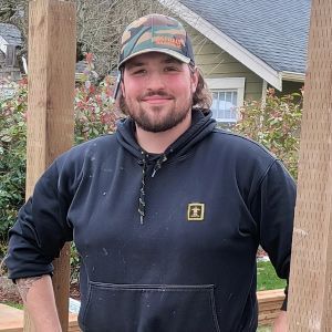 Person in a black hoodie and safety harness standing on a ladder outdoors, holding a tool belt.