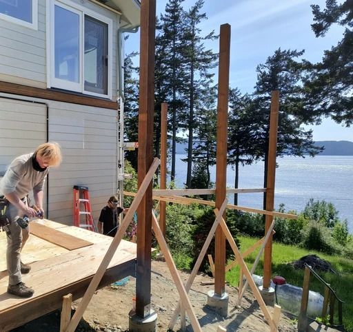 People building a wooden deck beside a lakeside house with tall support posts and tools nearby