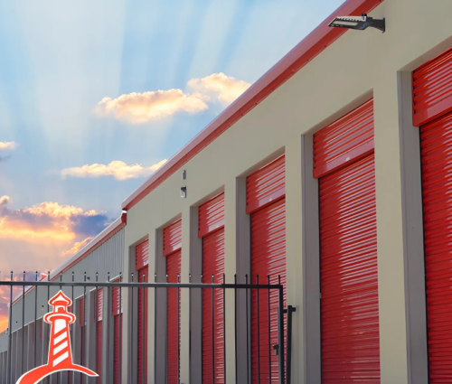 Row of storage units with red doors beside a black fence under a pastel sunset sky