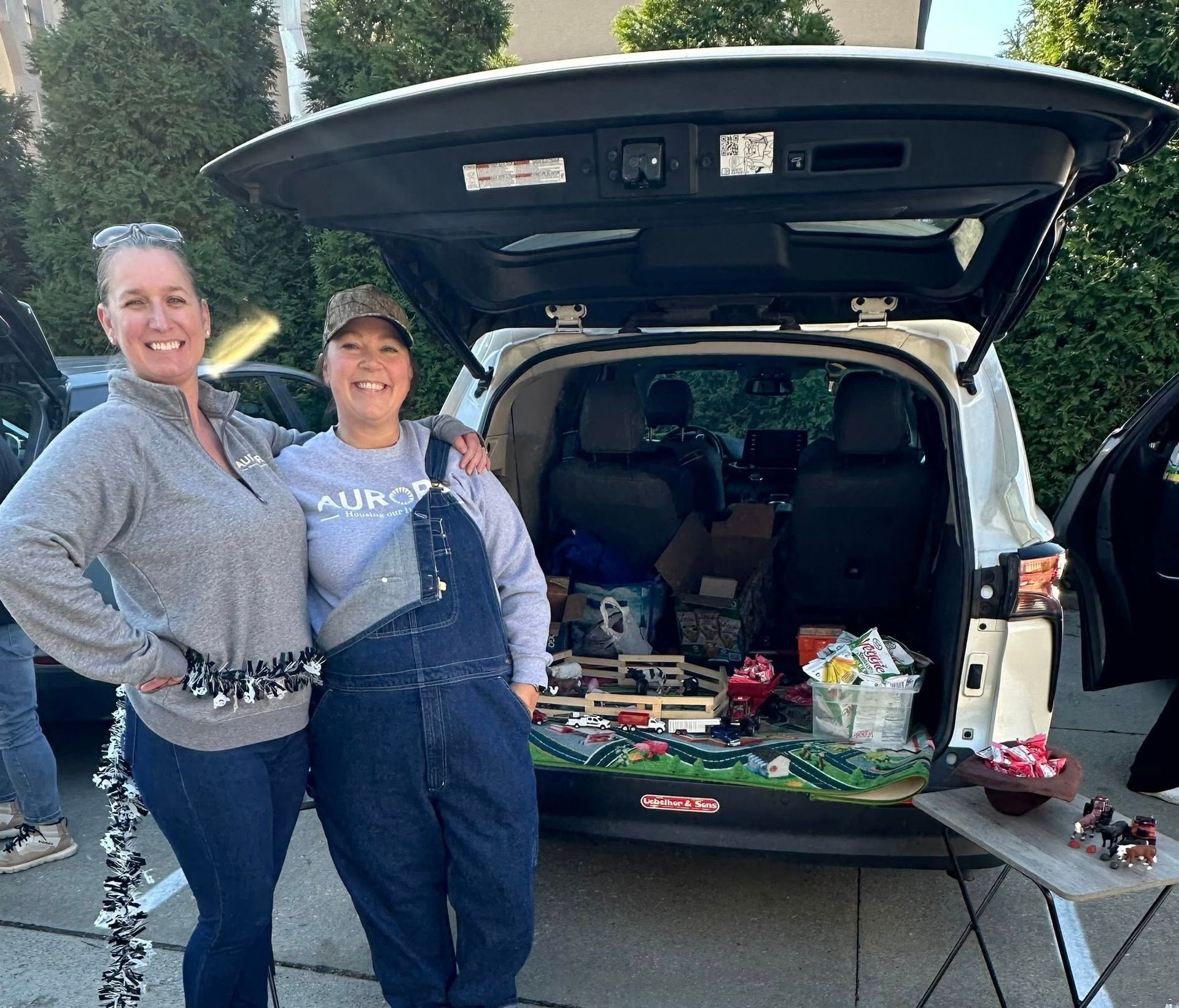 Two smiling people stand in front of an open vehicle trunk containing decorations and snacks for a trunk-or-treat event.