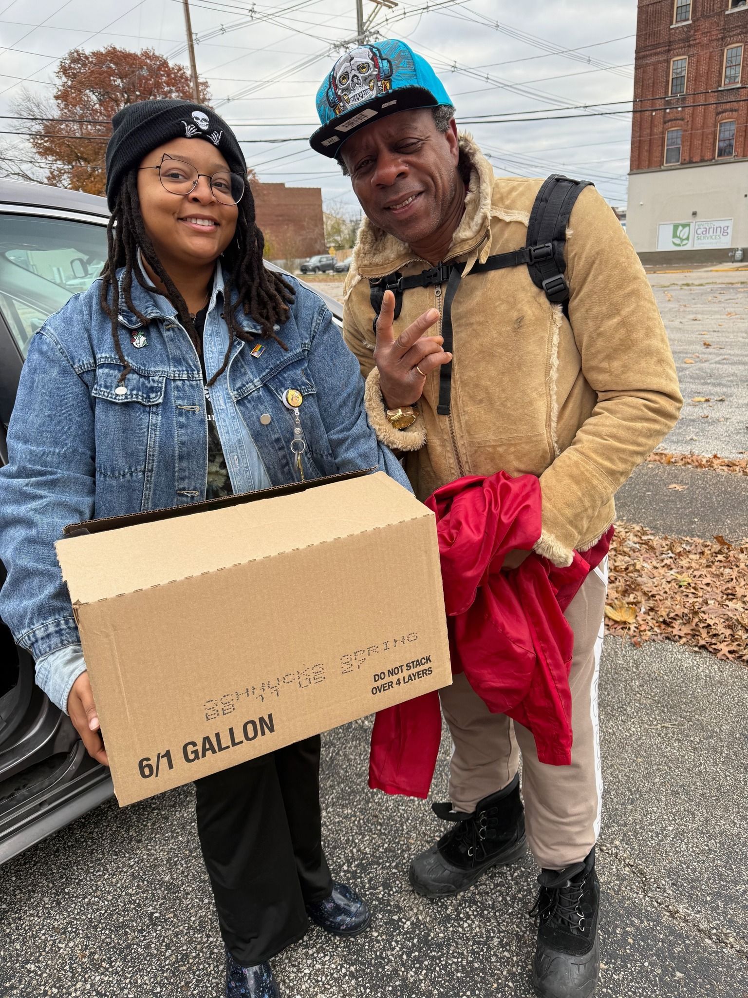 Two people smile outdoors, one holding a box labeled 