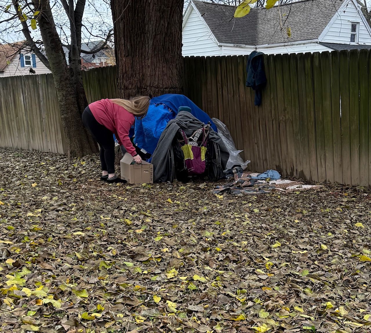 A person in a red top reaches into a cardboard box next to a makeshift shelter of blue tarps against a wooden fence.