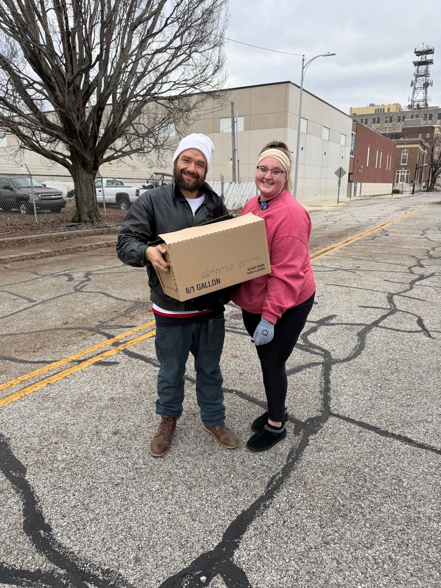Two people stand on a cracked asphalt road, smiling and holding a cardboard box between them, with a building behind them.