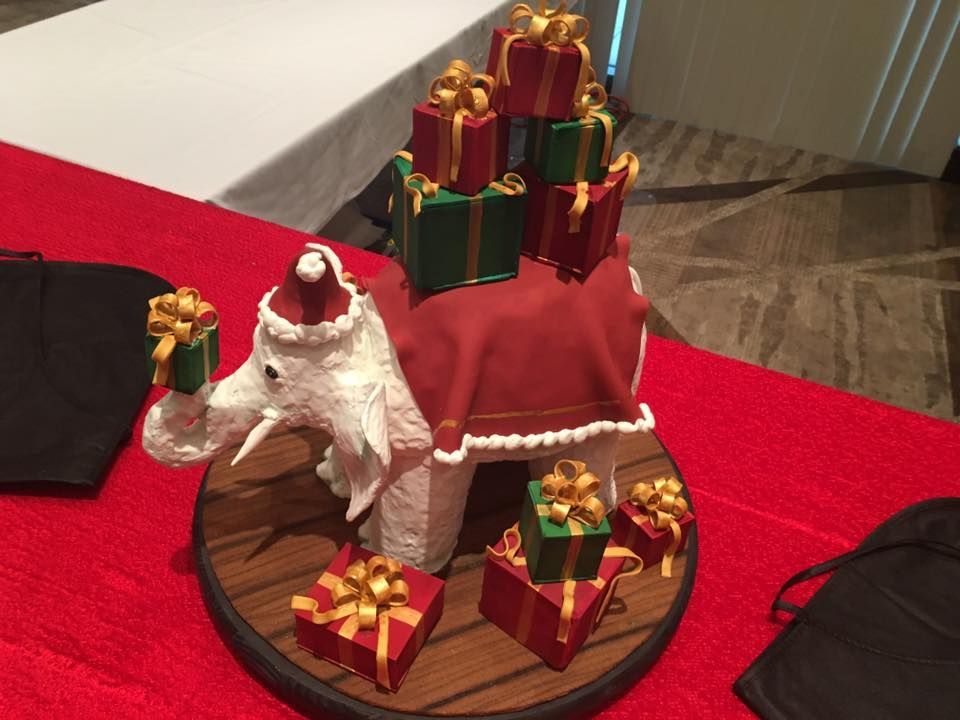 A white elephant figurine wearing a Santa hat and red blanket, surrounded by and carrying small, gift-wrapped presents.