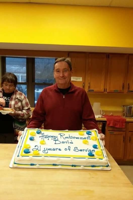 A man stands behind a white sheet cake with blue and yellow icing that reads, 