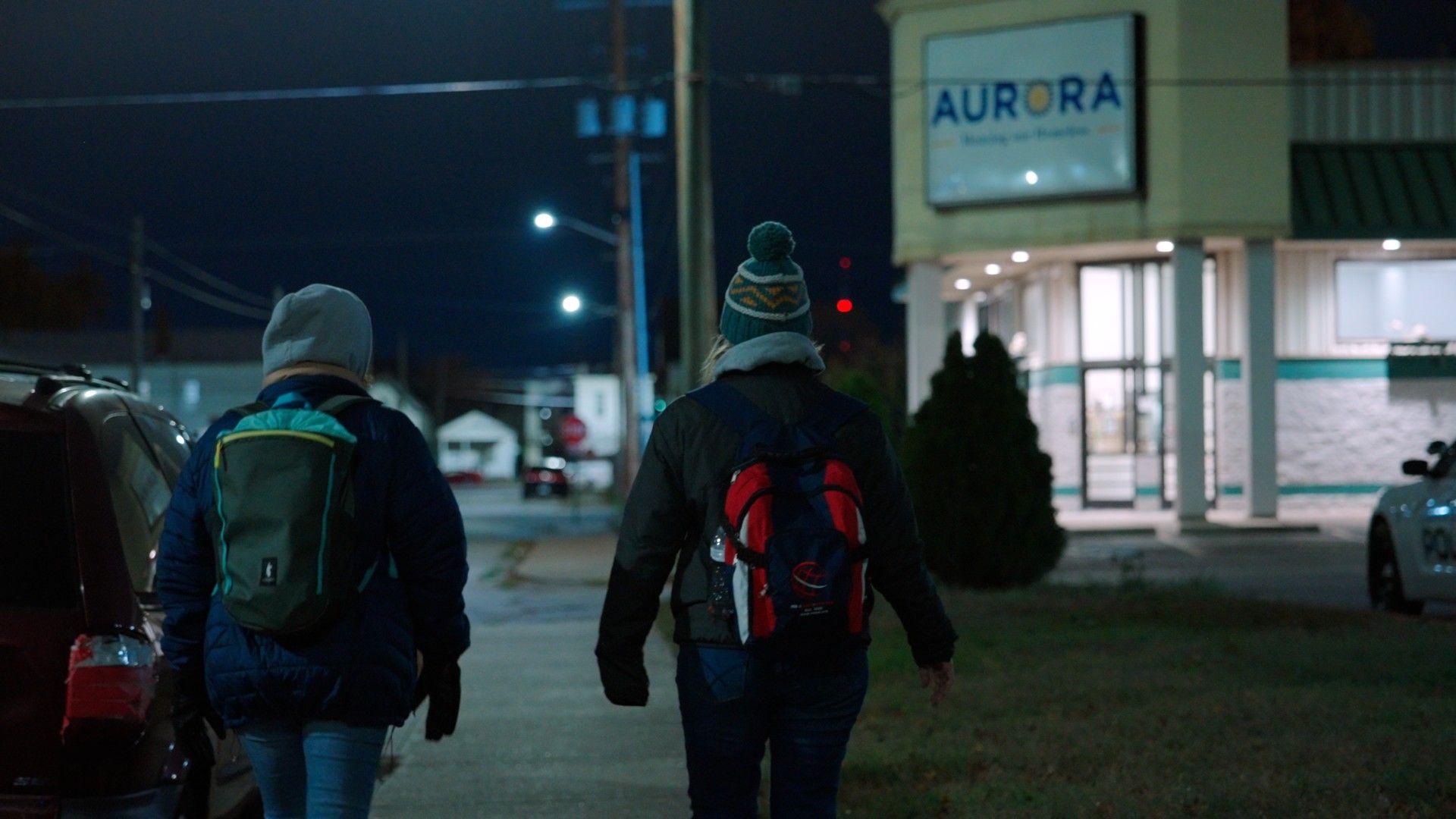 Two people with backpacks walk along a sidewalk at night past an Aurora building sign.