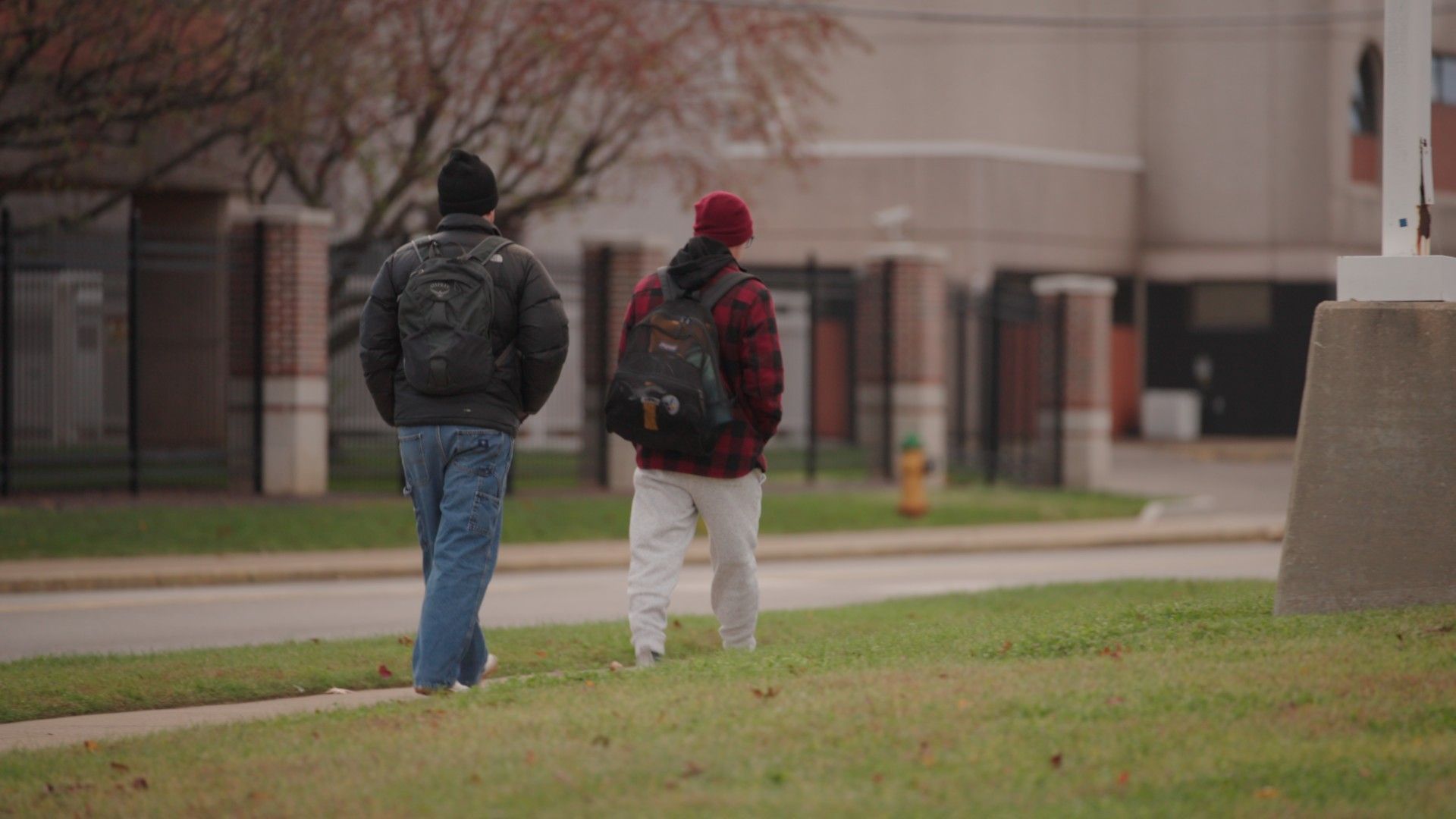 Two people with backpacks walking side-by-side along a sidewalk near a building on an overcast day.