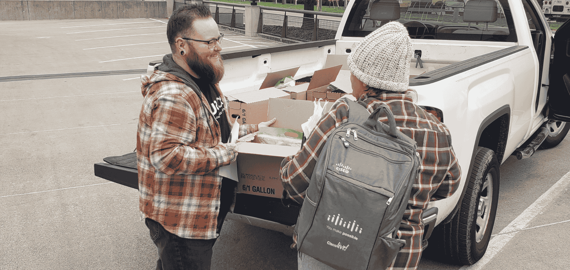 Two people in flannel shirts stand by an open truck bed, moving cardboard boxes filled with supplies.