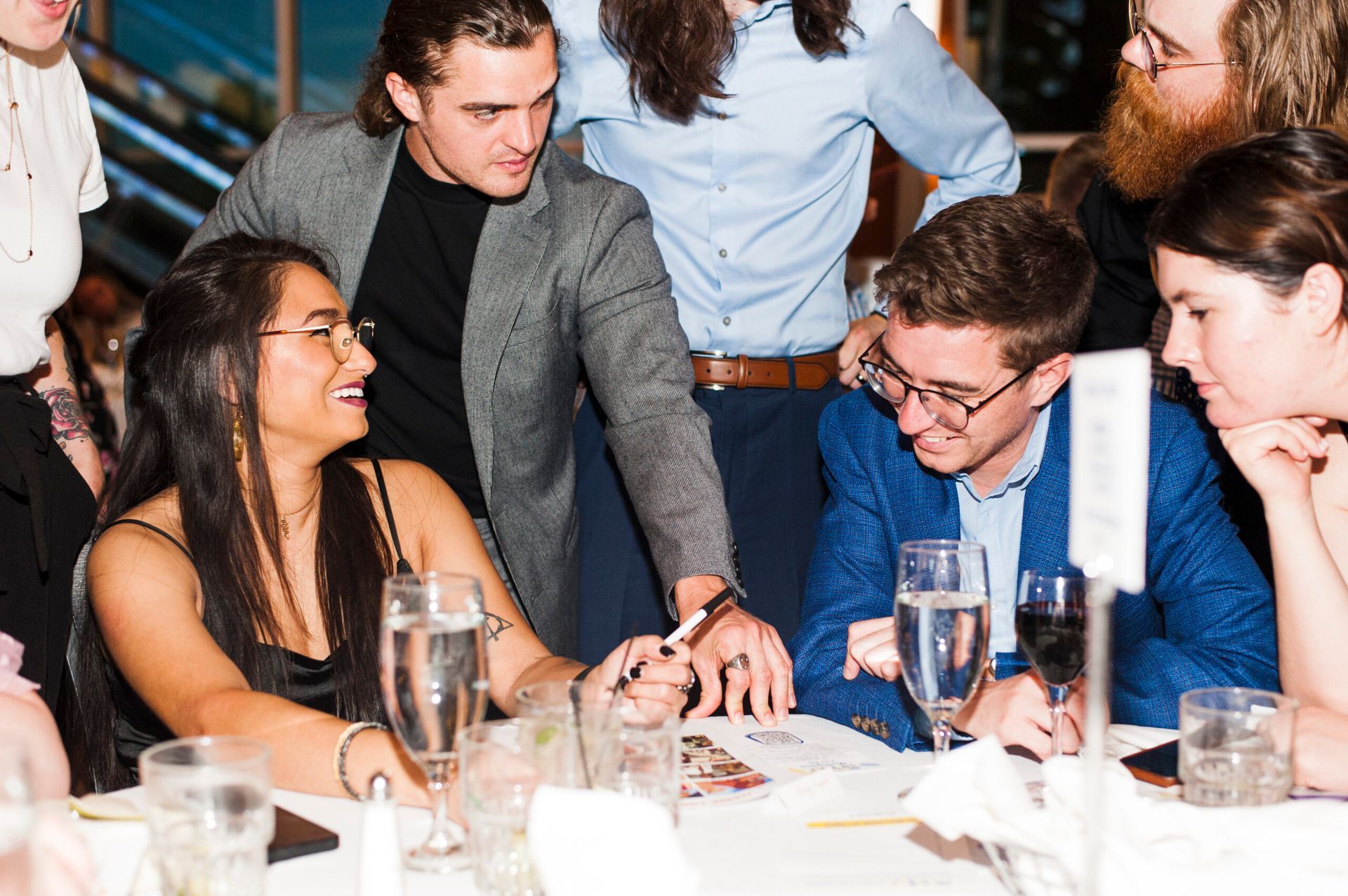 A group of people sitting and standing around a table at a formal event, looking at a document and smiling together.