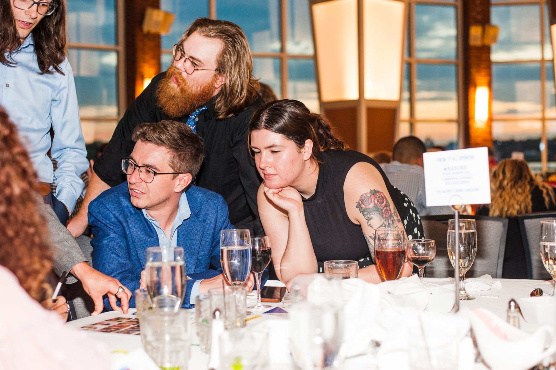 Four people gather around a dinner table at an evening event, leaning in to look at a document together.