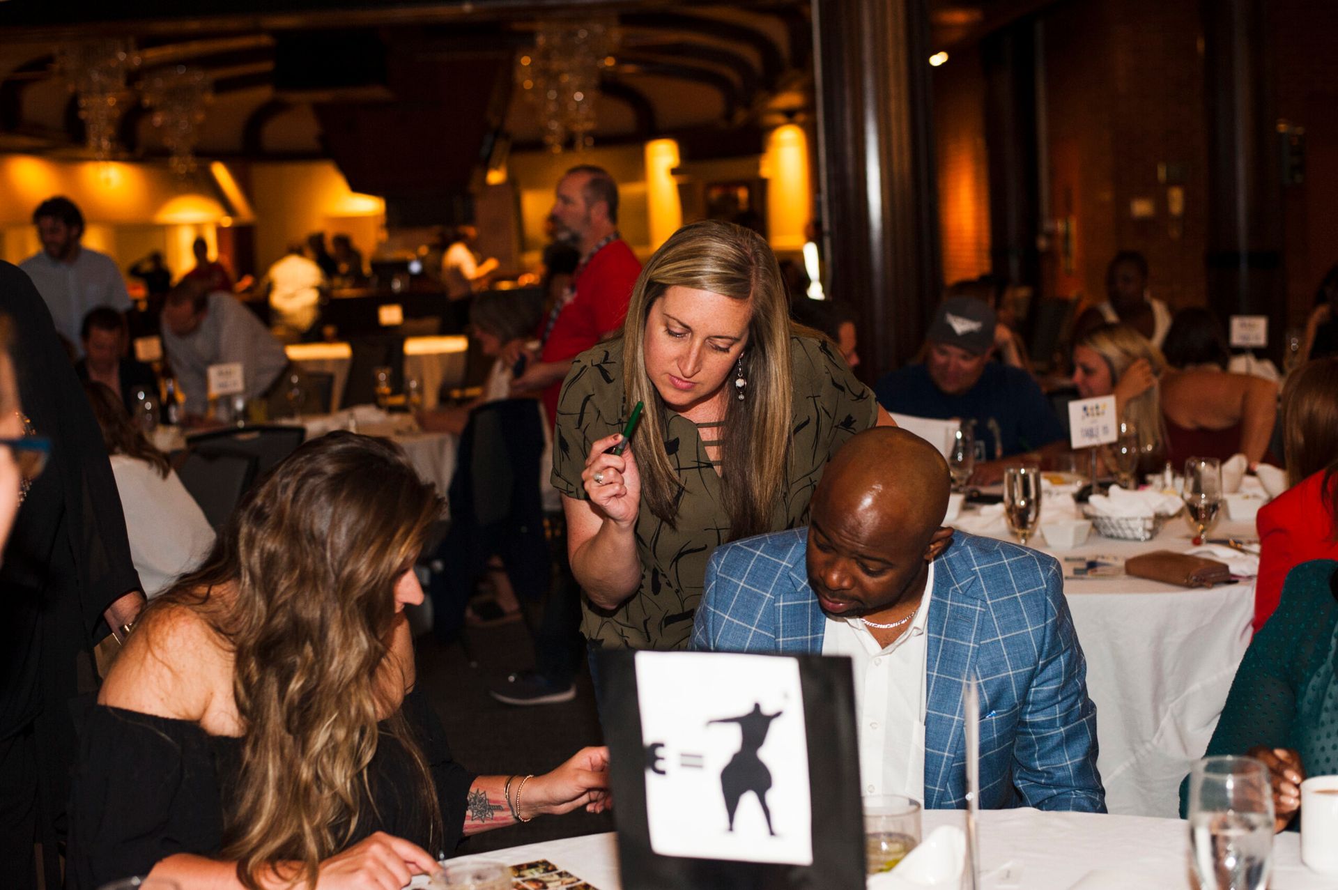 People seated at tables in a banquet hall, with a woman leaning over to show a printed graphic to a man in a blue jacket.