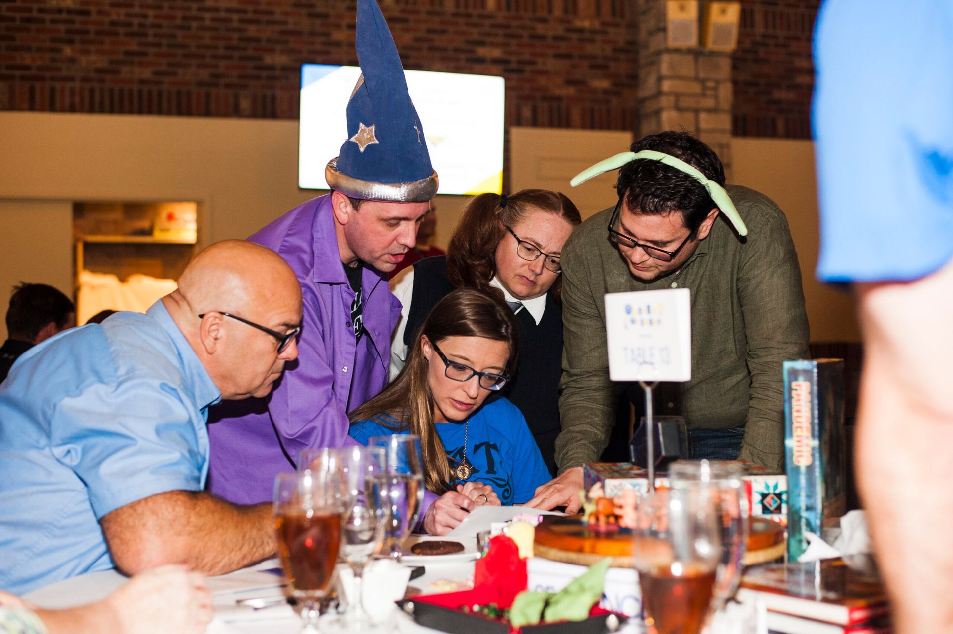 A group of people wearing costumes and glasses gather around a table to participate in a trivia game in a brick room.