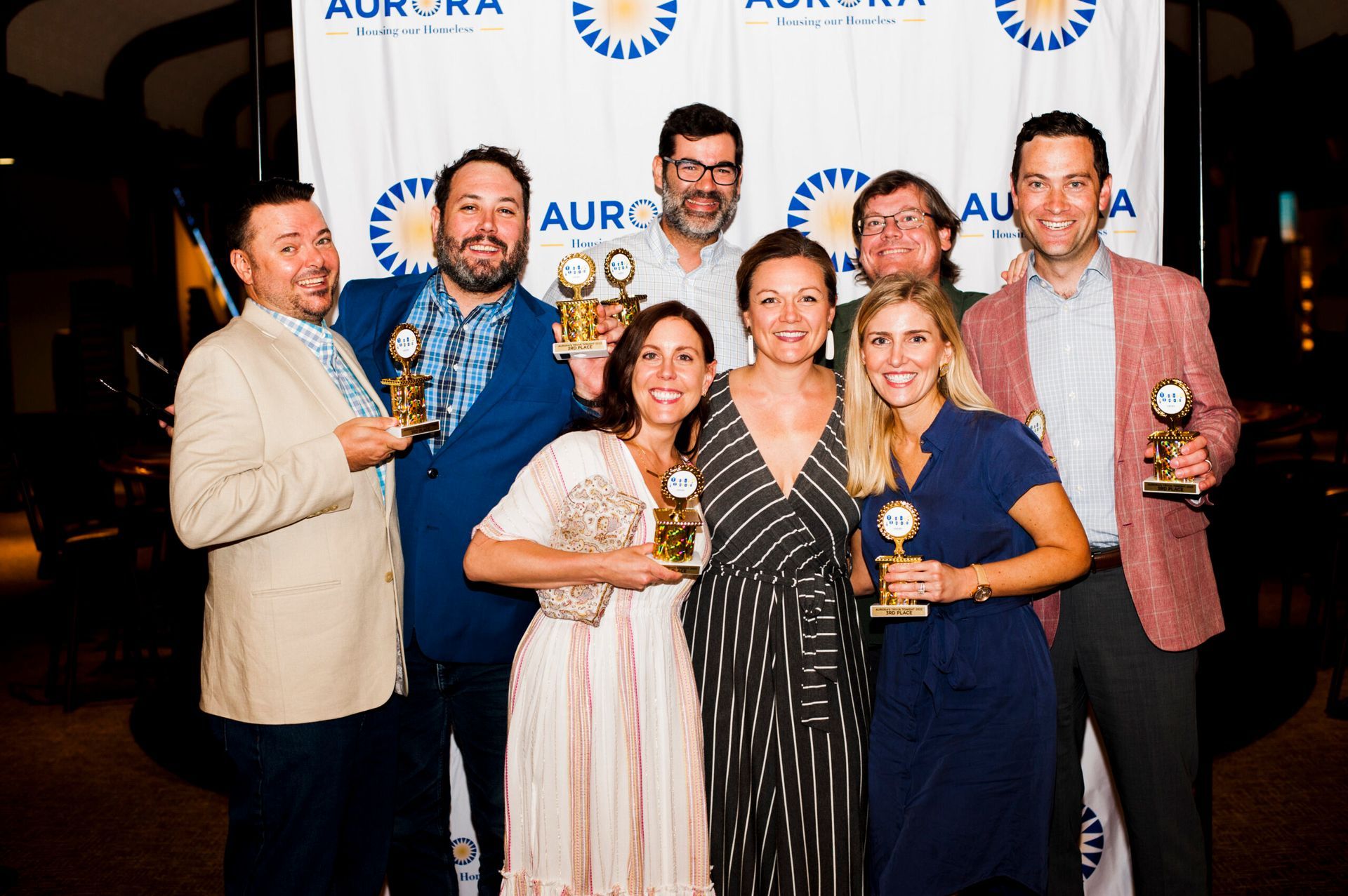 A group of eight people smiling while holding award trophies in front of an Aurora branded backdrop.