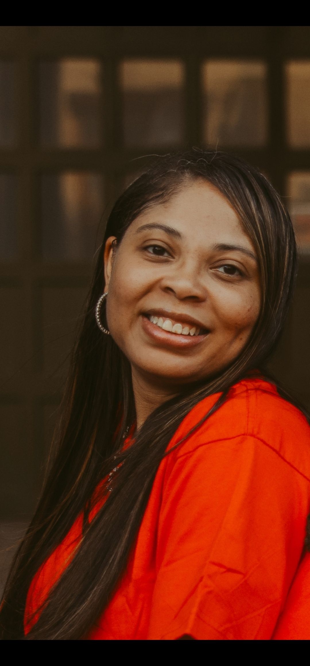 A smiling person with long dark hair wears a bright orange top and hoop earrings in a softly lit, warm-toned setting.