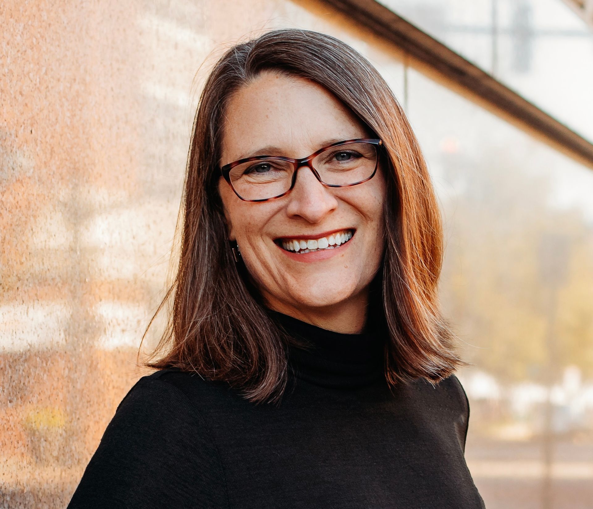 A person with shoulder-length brown hair wearing glasses and a black turtleneck, smiling in front of a textured wall.