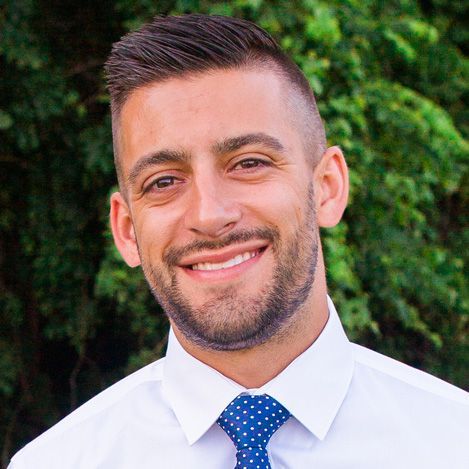 A smiling person with a close-cropped haircut, wearing a white collared shirt and blue polka-dot tie, outdoors.