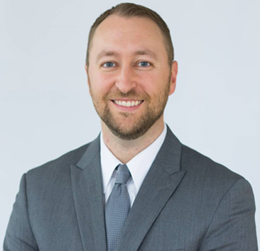 Professional headshot of a person wearing a grey suit and tie, smiling against a plain light background.