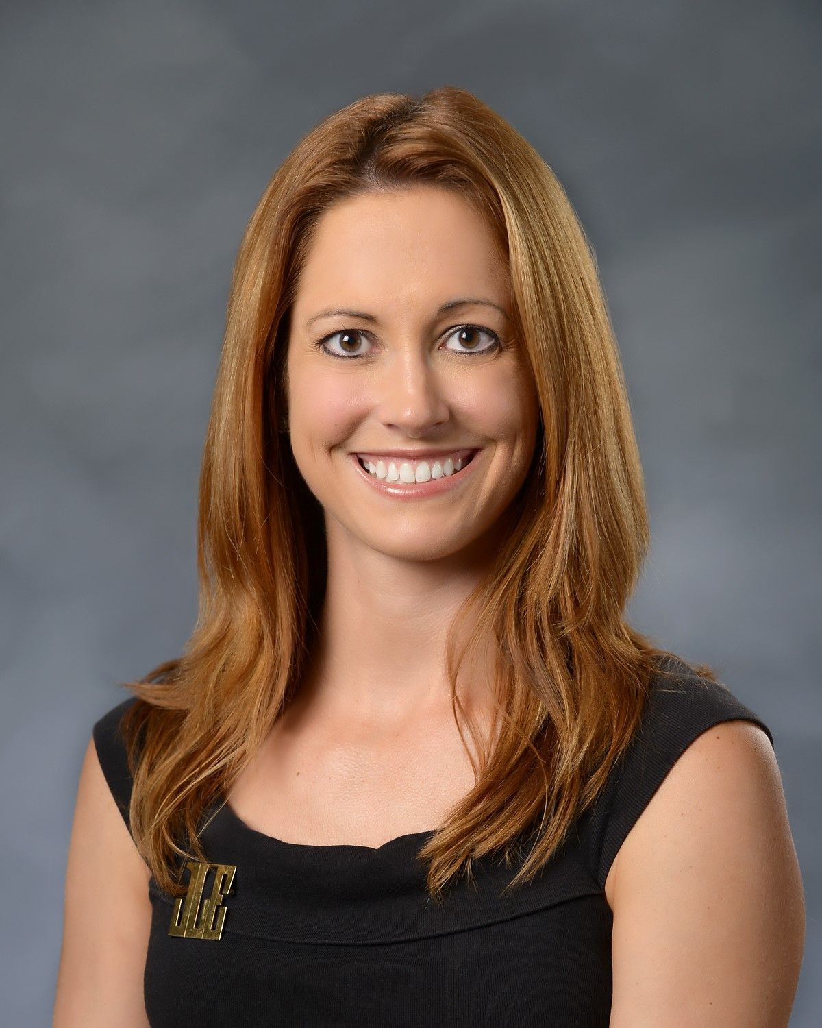 A person with shoulder-length light brown hair smiling at the camera, wearing a dark top, against a gray background.