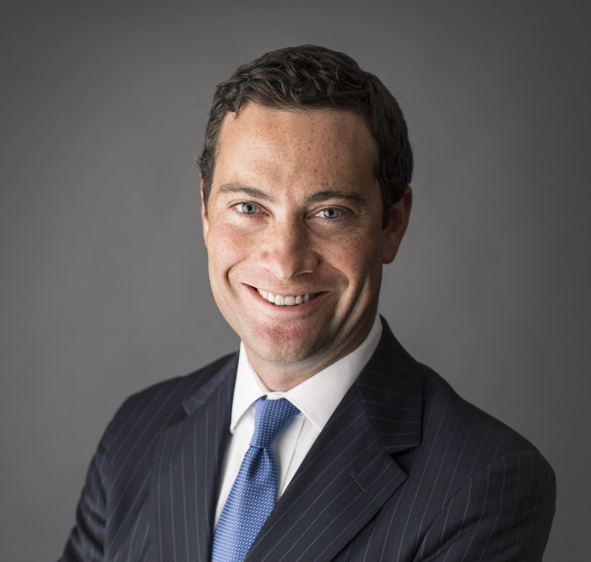 A smiling person in a blue pinstripe suit, white shirt, and blue patterned tie, set against a solid gray background.