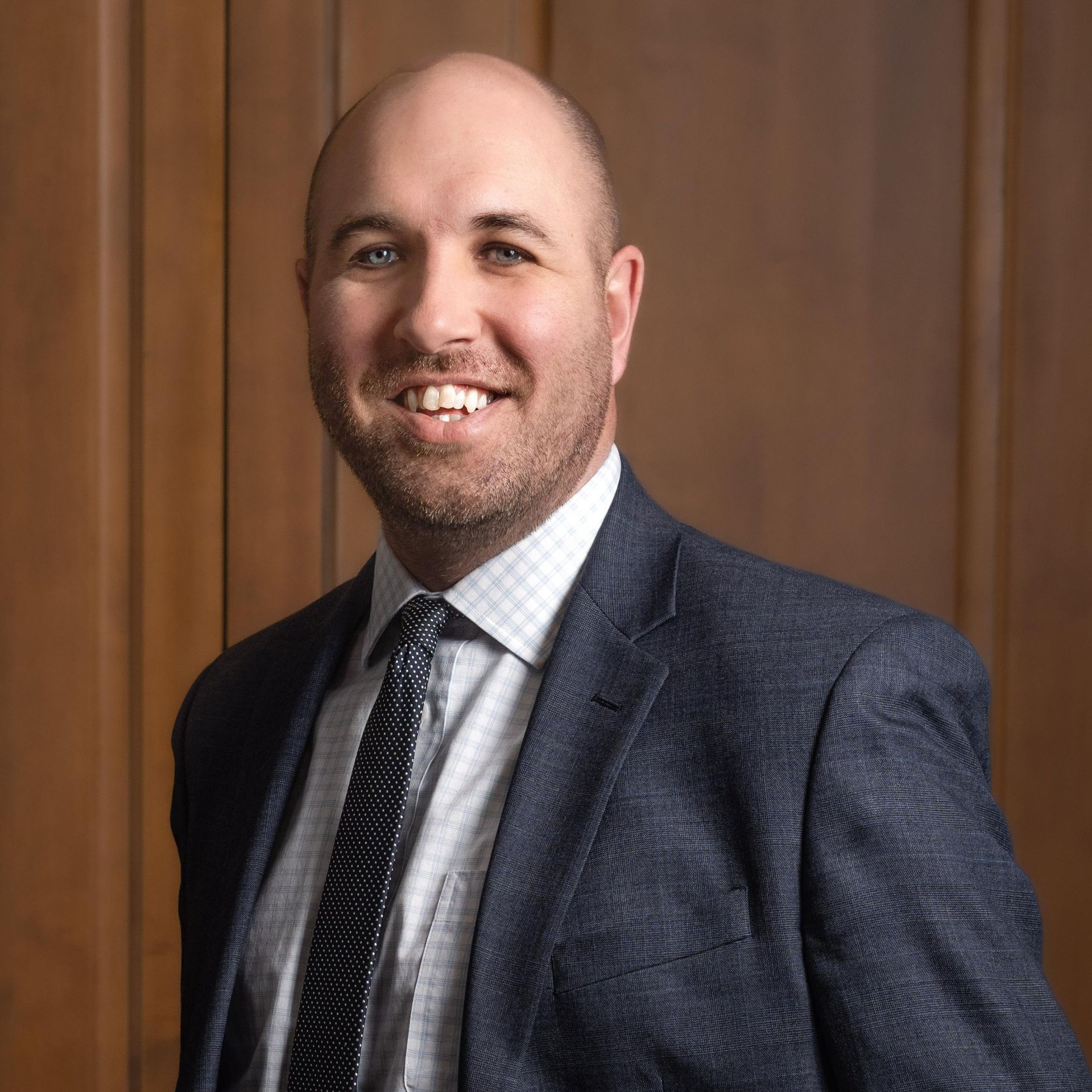 A person in a blue textured blazer, light button-down shirt, and patterned tie smiling against a wood-paneled background.