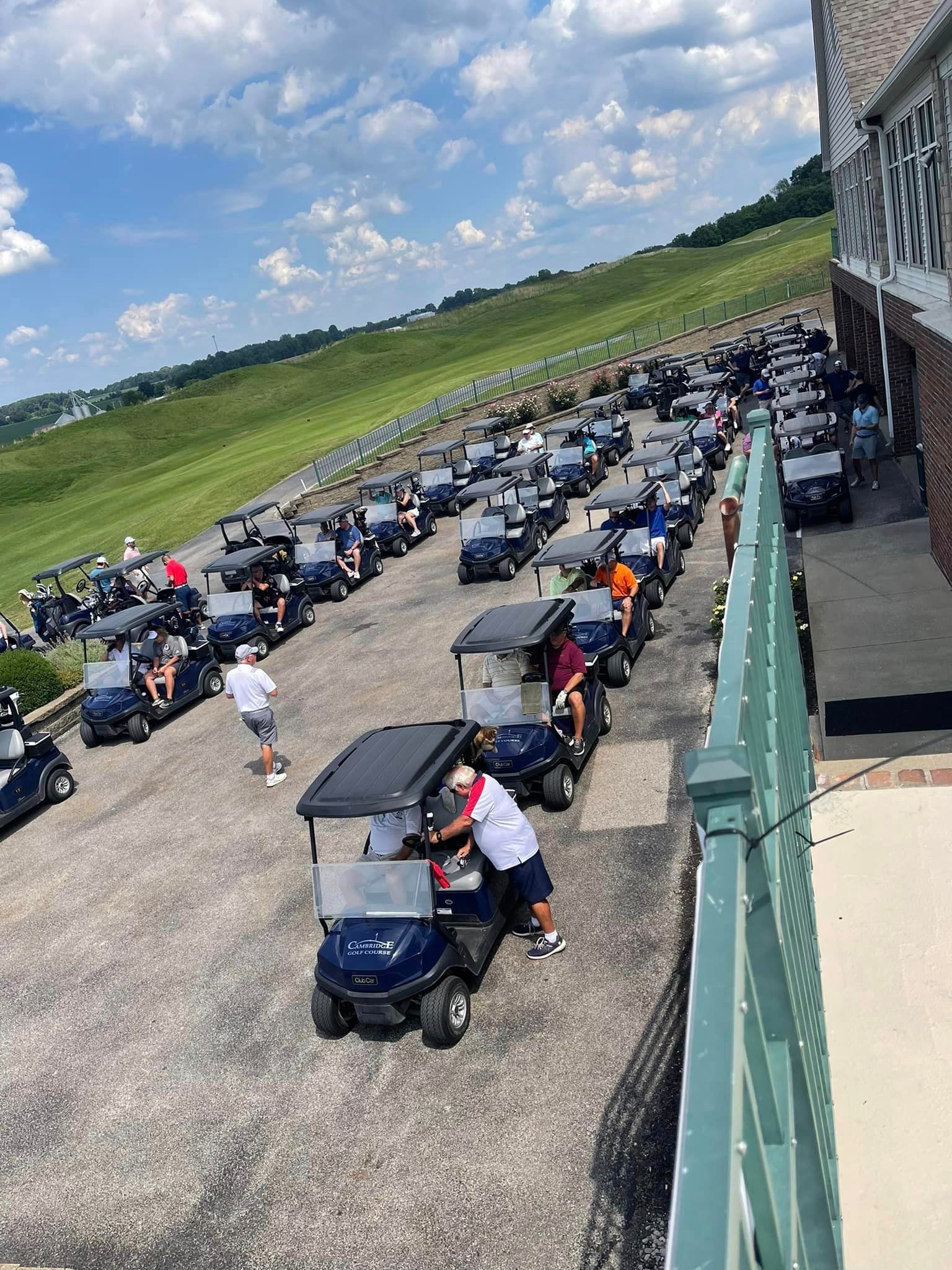 A large group of golf carts is lined up on a gravel path next to a building on a sunny day.