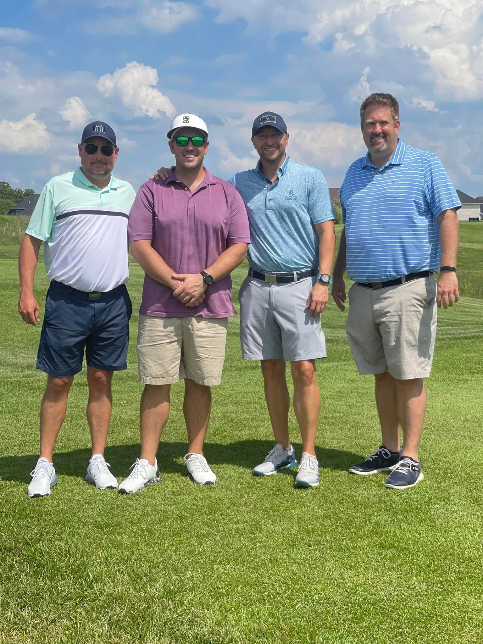 Four men stand in a sunny, grassy field, smiling and wearing golf attire, posed for a group photo.
