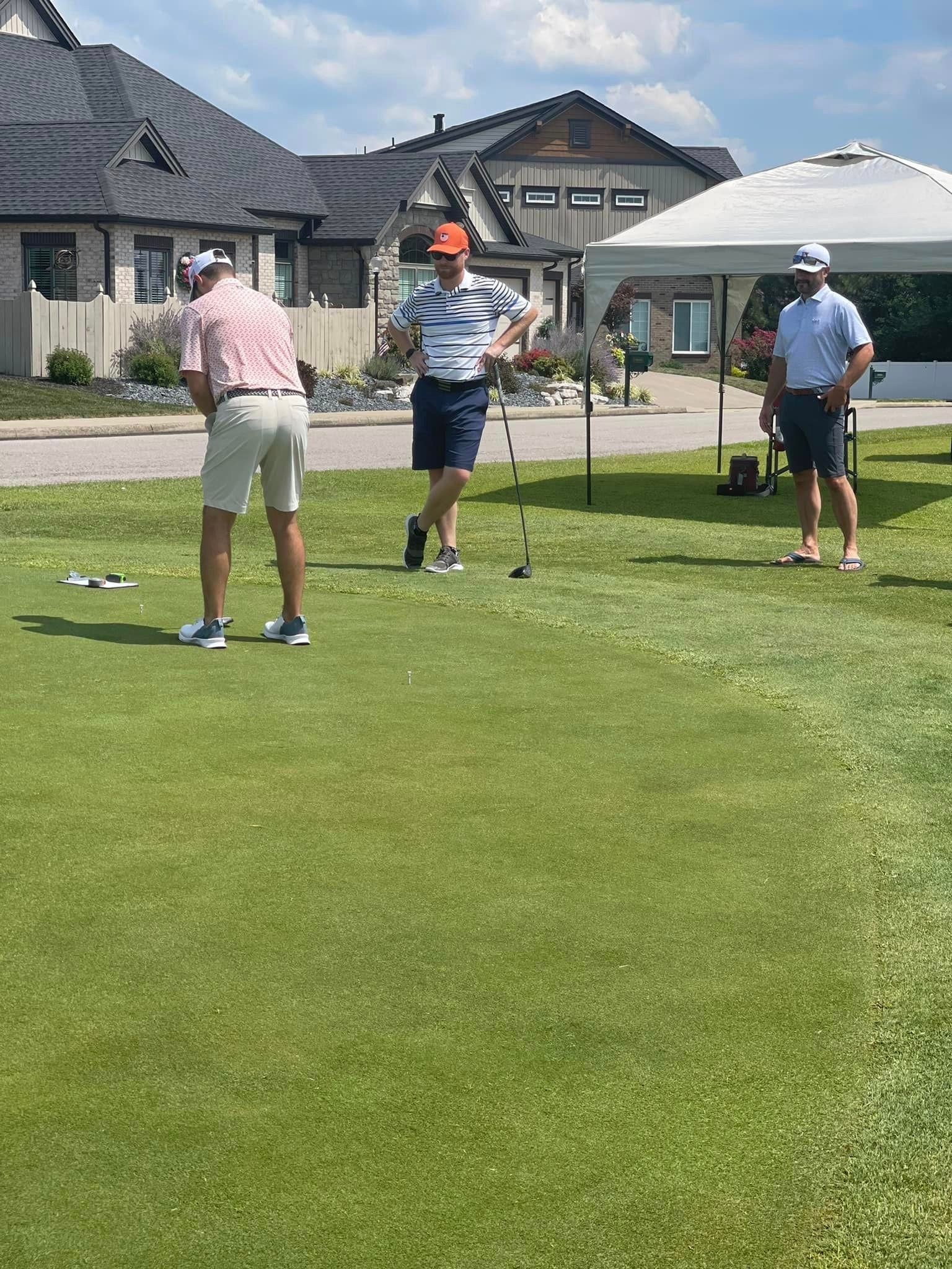 Three people wearing golf attire stand on a putting green in front of a clubhouse with a white canopy tent nearby.
