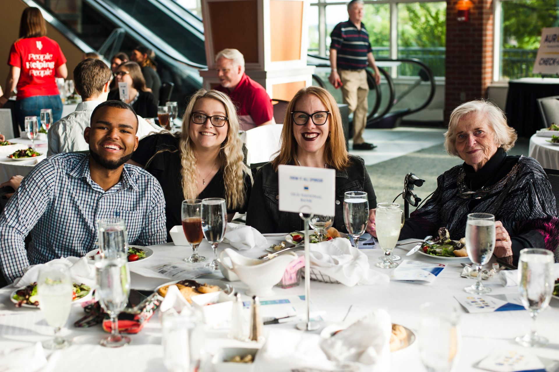 Four people smiling at a formal table setting with food and drinks in a bright, indoor event space.
