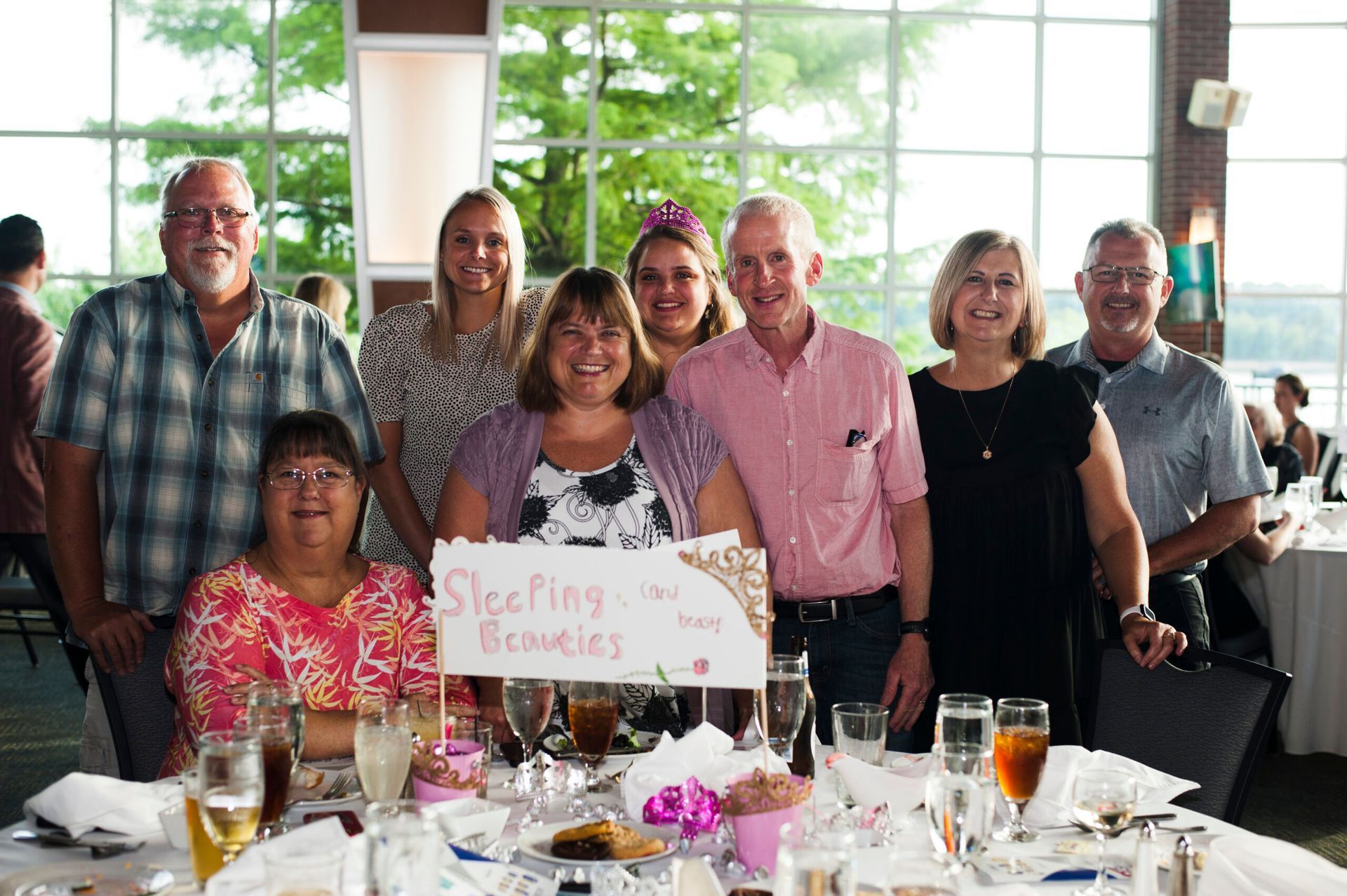 A group of nine people smiling together at a dining table at an indoor event, with a sign reading