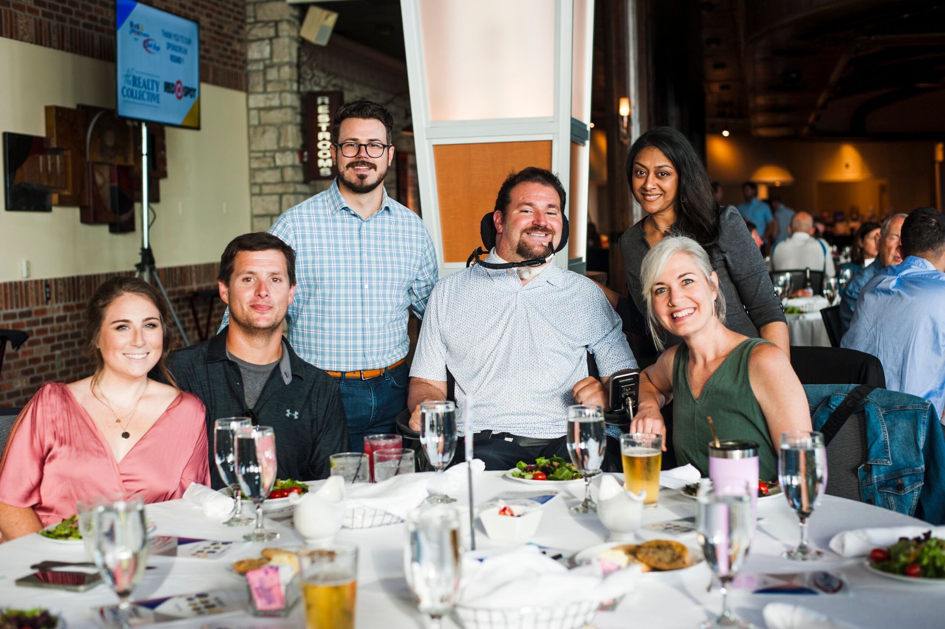 A group of six people posing for a photo at a dining table in a warmly lit, casual restaurant setting.