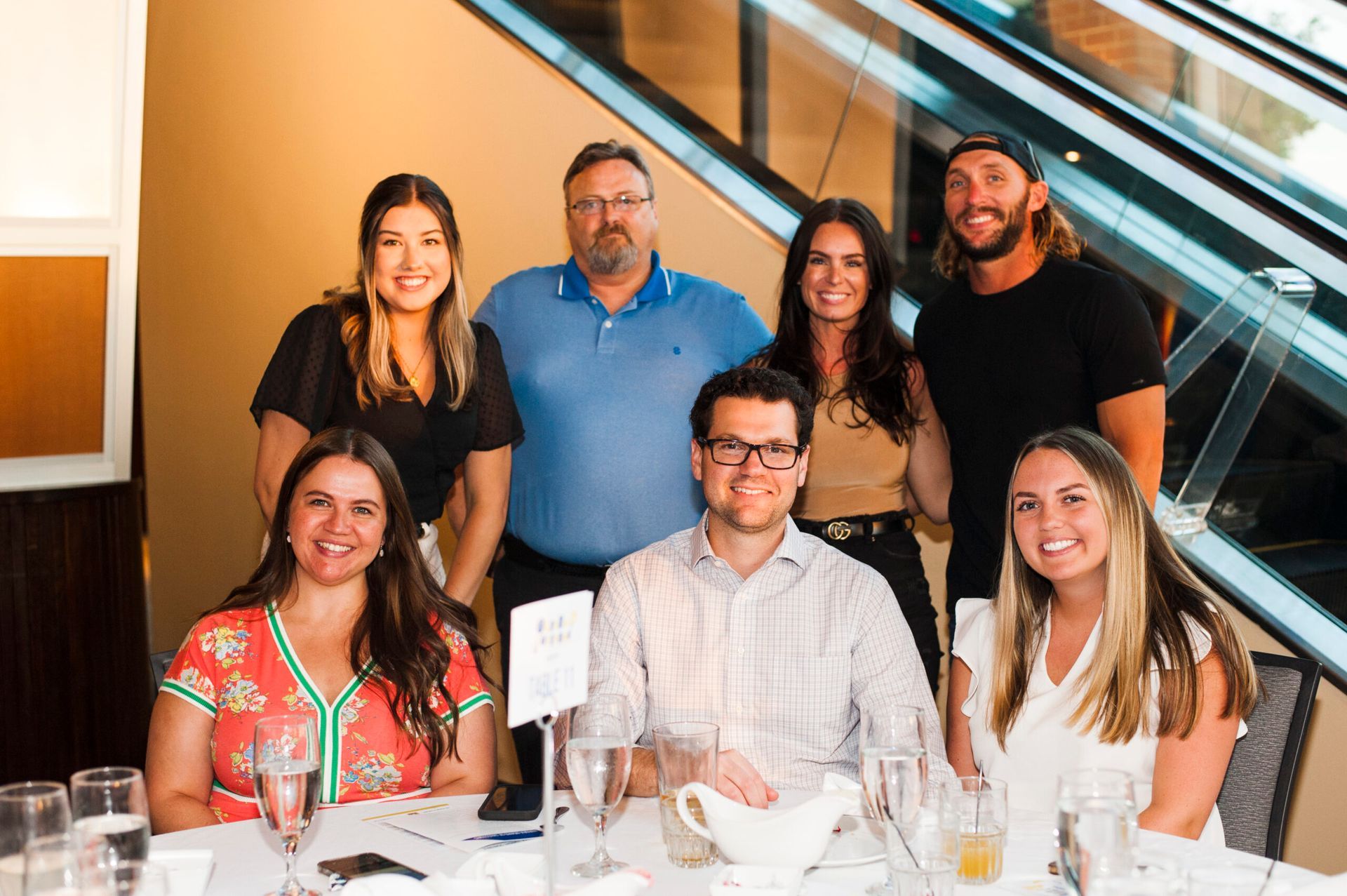 A group of seven people smiling for a photo around a table at an indoor event with an escalator in the background.
