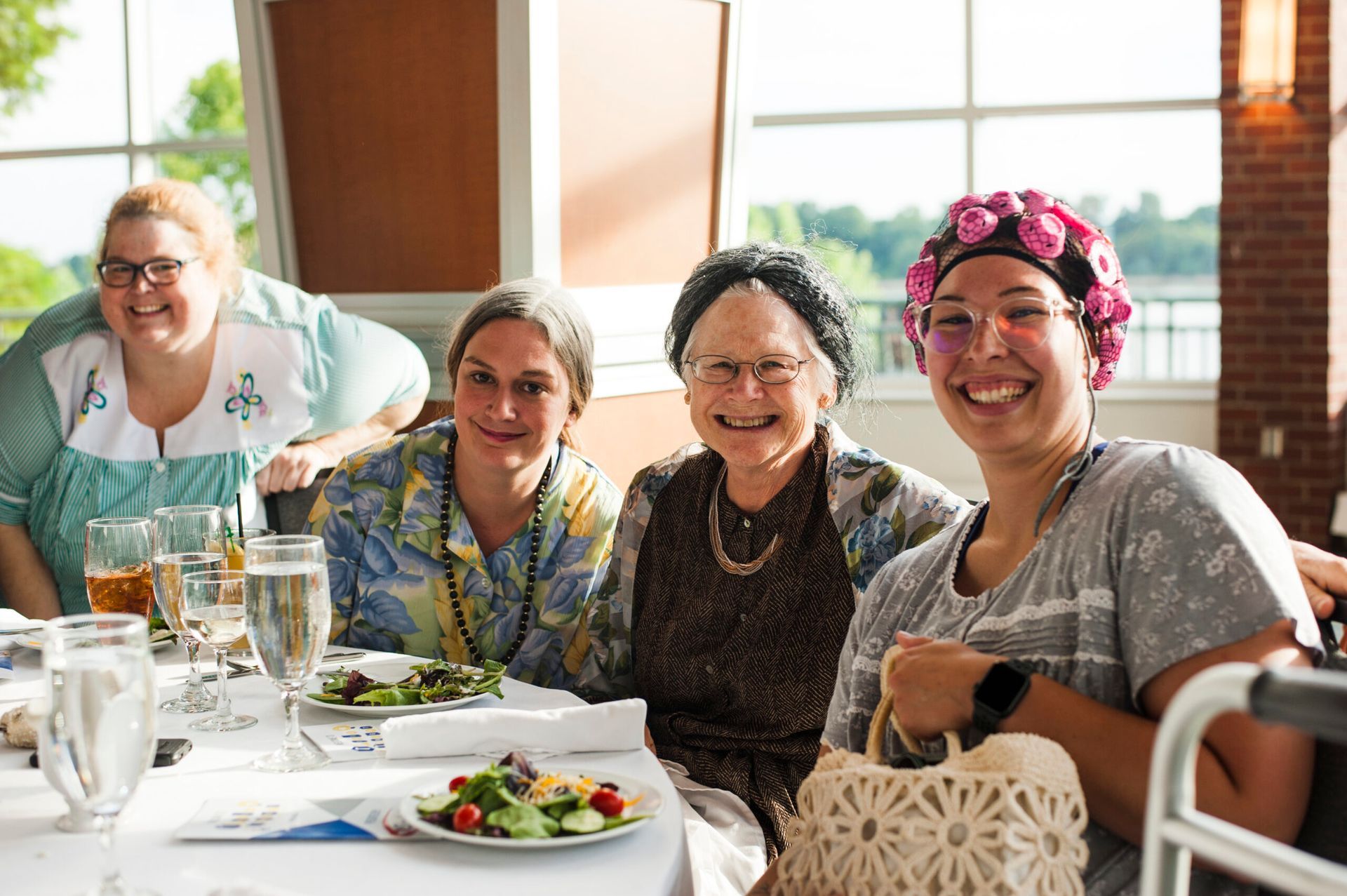 Four people dressed in costumes with hair curlers or head coverings smile while sitting at a table set for a meal.